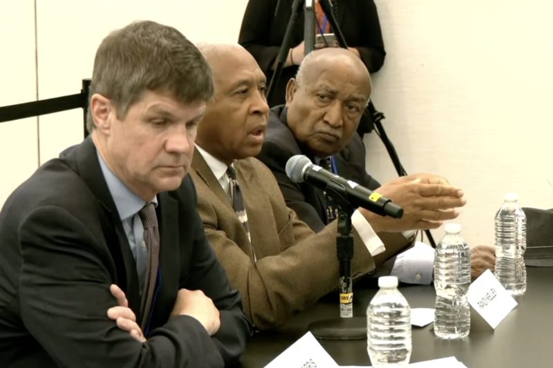 Three men sitting at a table. One is speaking into a microphone.