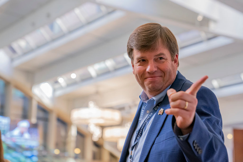 A man in a blue shirt and jacket smiling and pointing.