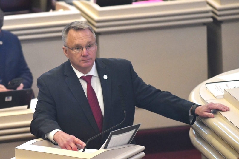 A man in a dark blue suit and a red tie standing at a lectern while resting his hand on a nearby desk.