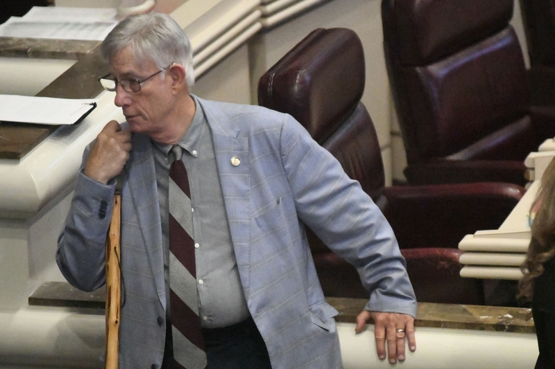 A man in a blue jacket, gray shirt and black and gray striped tie leaning on a walking stick.
