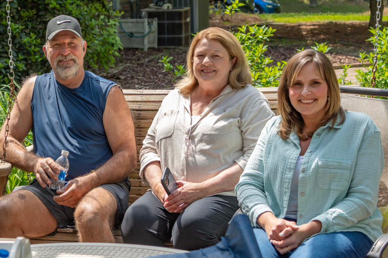 A man in a baseball cap and sleeveless T-shirt sitting next to two women in button up T-shirts on a bench in a backyard.