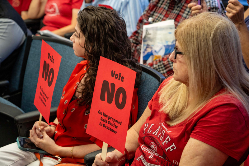 Two women in red T-shirts holding signs saying Vote No on the proposed data center in Bessemer