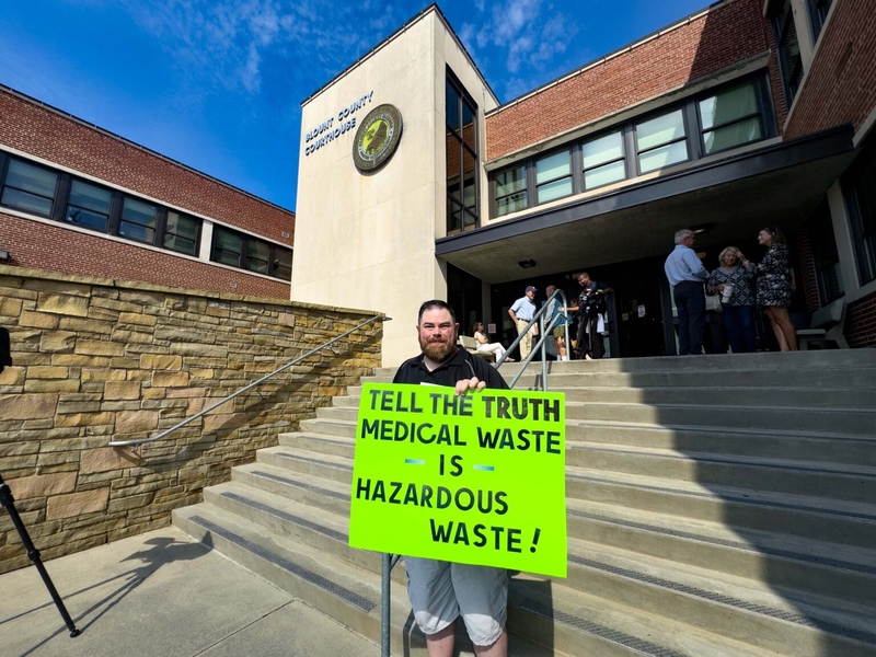 A man holding a green sign saying Tell the Truth: Medical waste is hazardous waste