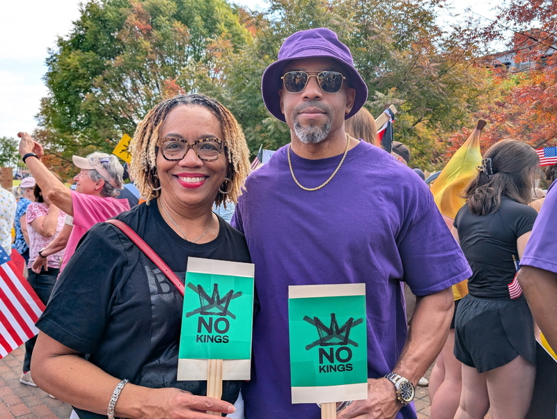 A woman in a black T-shirt and a man in a purple T-shirt and hat holding signs saying No Kings