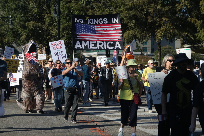 A crowd of people marching with a large banner saying "No Kings In America"