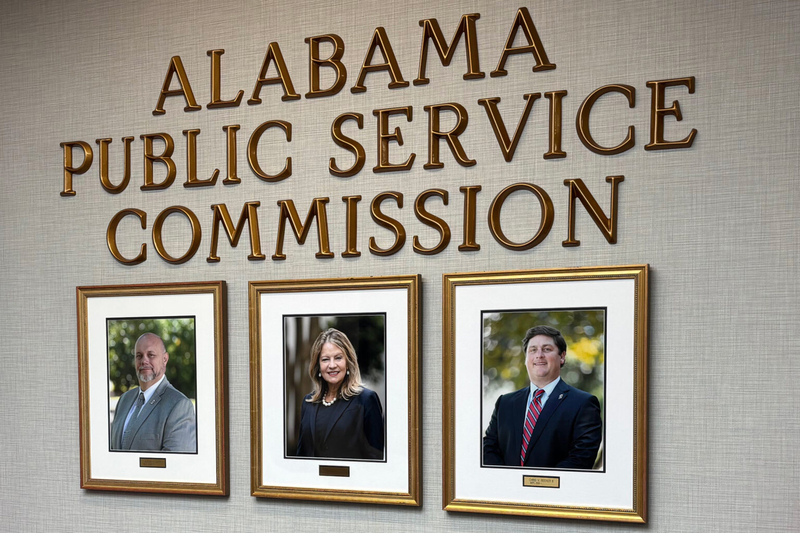 Letters spelling out Alabama Public Service Commission on a wall. Below, two framed portraits of men in business suits flanking a woman in a business suit.
