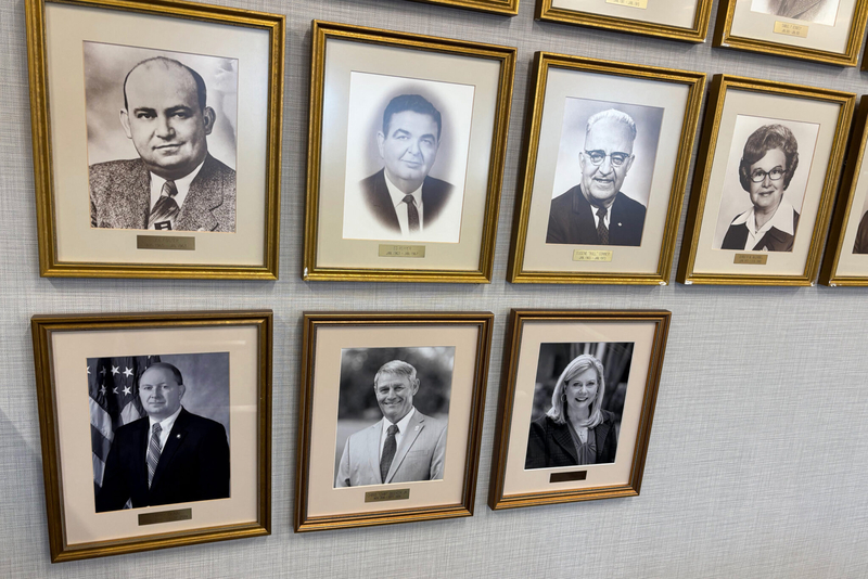 Seven framed black and white portraits of six men and one woman in business suits on a wall
