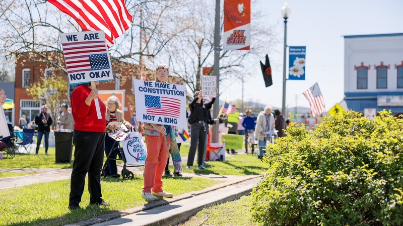 A group of protestors holding signs saying We Are not Okay and We have a constitution not a king in a town square.