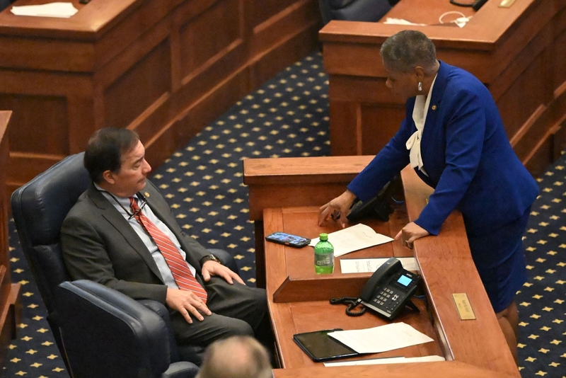 A woman in a blue dress speaking to a seated man ina charcoal suit