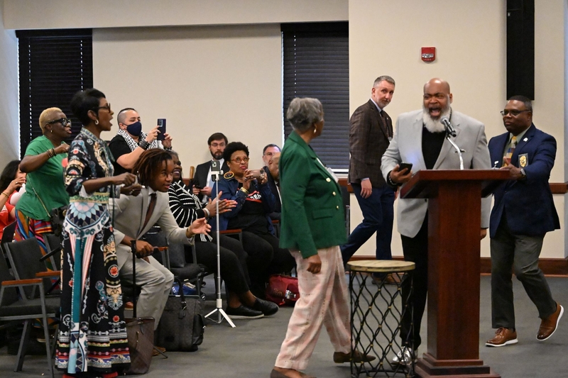 A man in a gray jacket speaking as people protest around him