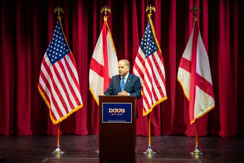 A man behind a lectern saying Doug For Alabama with American and Alabama flags behind him