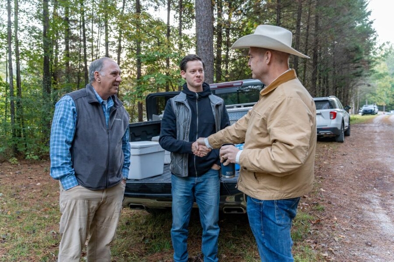 Two men in vests speaking with a man in a barn jacket and hat