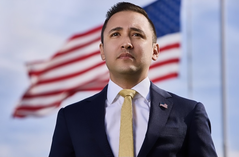 A man in a dark suit jacket and yellow tie stands in front of an American flag