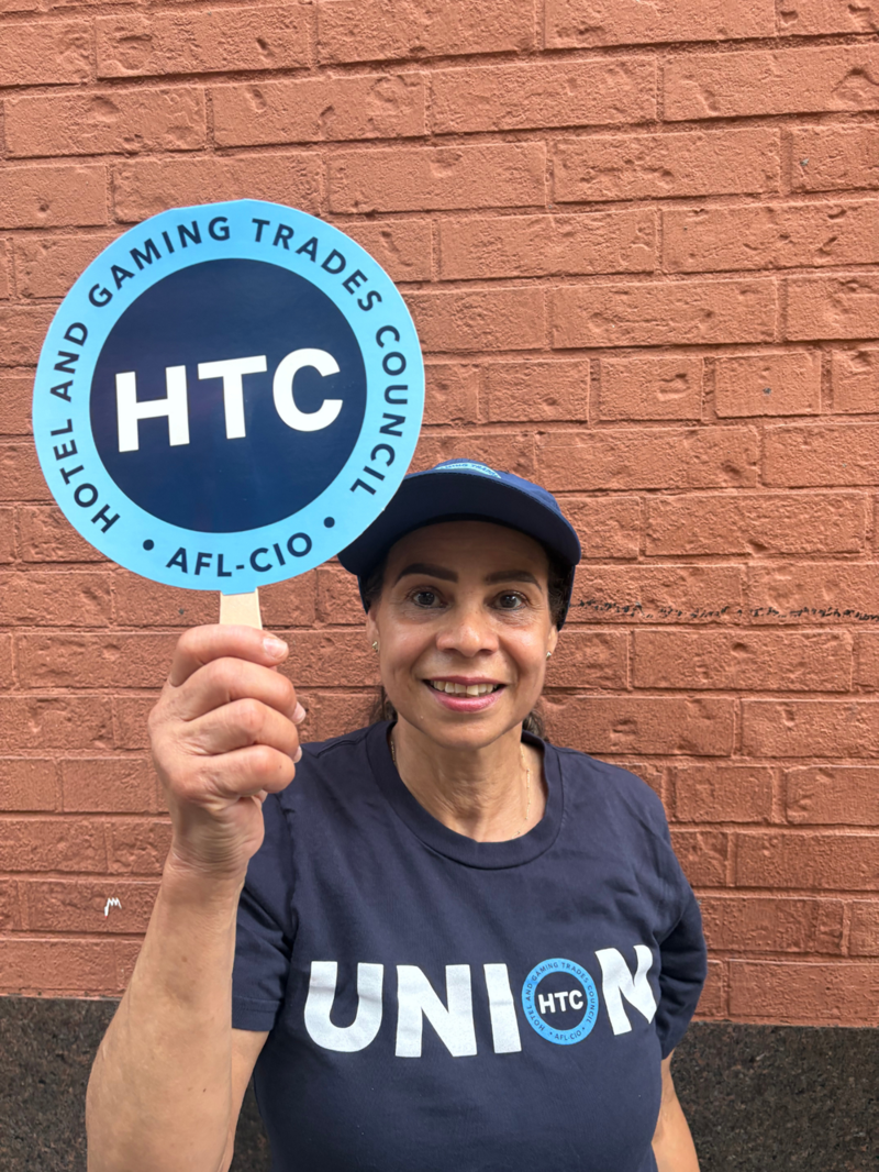 A photo of Arisnelda Rodriguez smiling and holding up a sign that reads, "HTC: Hotel and Gaming Trades Council" and wearing a shirt that says "Union."