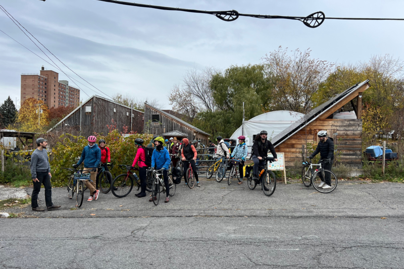12 or so cyclists have mounted their bicycles in front of some small wooden structures near the street.