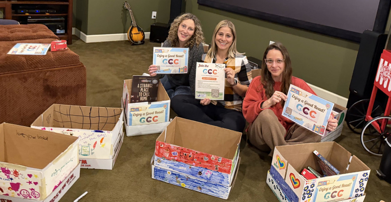 Three women hold signs saying "Enjoy a good read!" while packing books into boxes.
