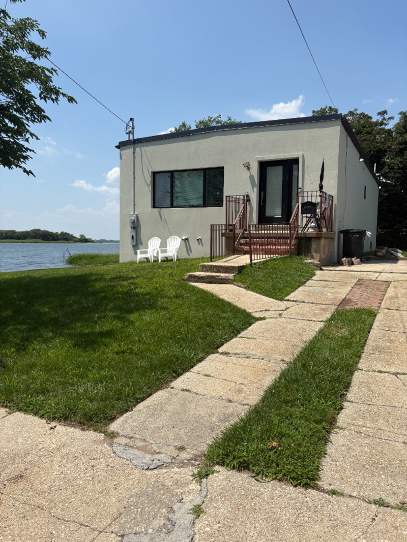 At the end of a long driveway near the water, a small single-family home with two plastic beach chairs out front.
