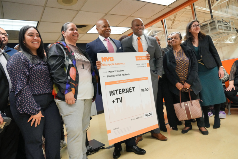 A photo of former New York City Mayor Eric Adams holding a large sign stating, "NYC Big Apple Connect. From: Mayor Eric Adams, To: 300,000 NYCHA Residents, Internet + TV = $0," standing in a line of six other people.