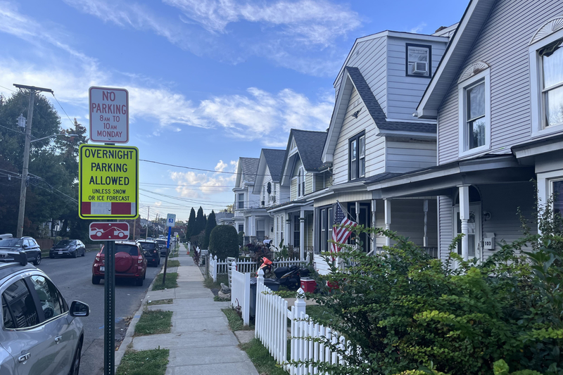 A perspective photo of a suburban-looking block with a row of two-story houses, a sidewalk, and the street with a few cars parked.