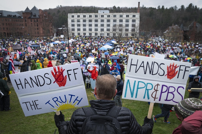 Crowd gathered at a protest. Person in foreground holds signs reading "Hands Off The Truth" and "Hands Off NPR &amp; PBS.