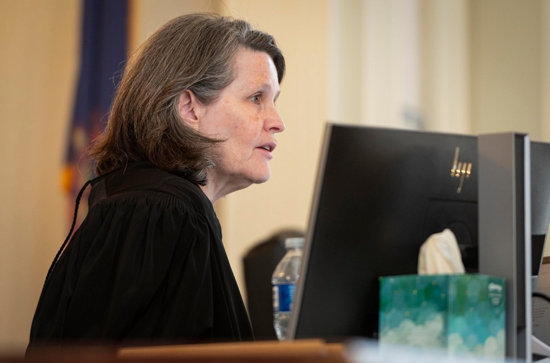 A judge in a black robe sits at a desk behind a computer monitor, with a water bottle and tissue box nearby, speaking in a courtroom setting.