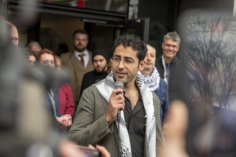 A man in glasses speaks into a microphone outside a building, surrounded by a group of people, some of whom are watching him attentively.