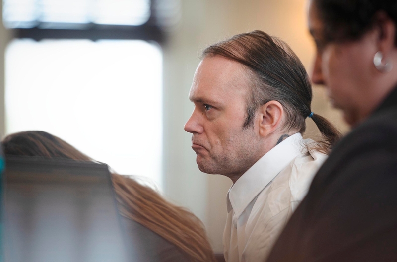 A man with a ponytail, wearing a white shirt, sits in a courtroom flanked by two other people.