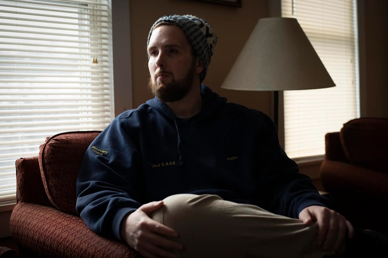 A man with a beard and knit hat sits on a sofa in a dimly lit room, with light coming through window blinds behind him.
