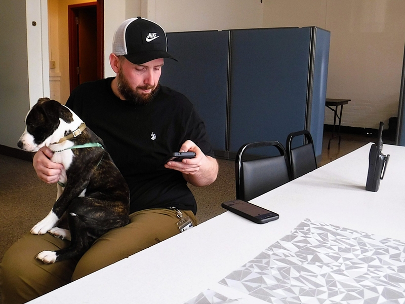 A man sitting at a table with a dog on his lap looks at his phone. Two smartphones and a walkie-talkie are on the table.