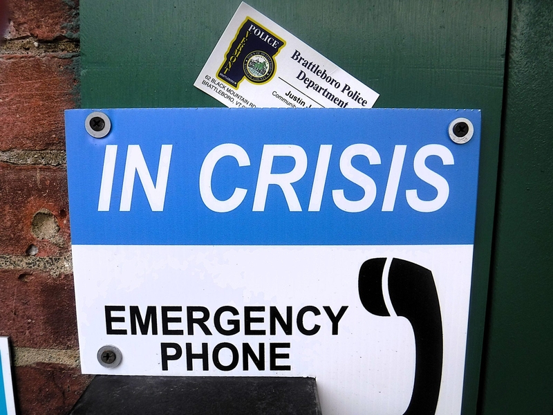 A sign reading "In Crisis Emergency Phone" is posted on a wall, with a Brattleboro Police Department business card tucked behind it.
