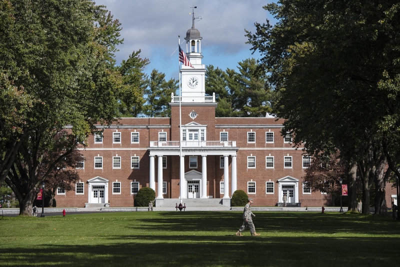 A traditional brick building with a central white clock tower, surrounded by lush green trees under a partly cloudy sky.