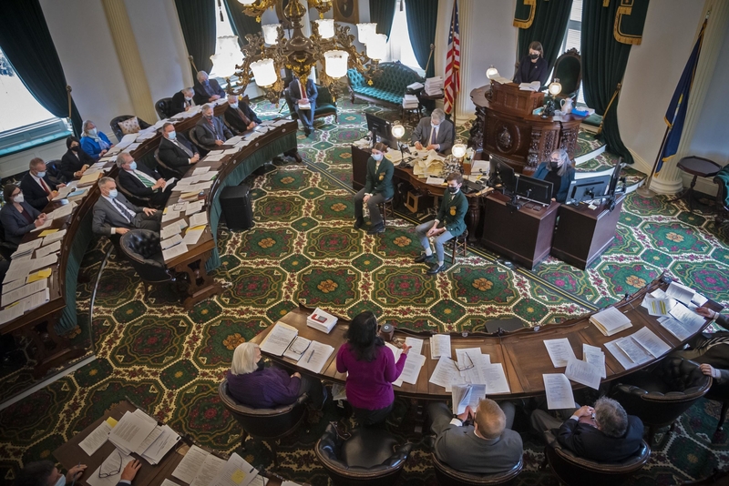 A legislative session in a large ornate chamber with several people seated around a horseshoe-shaped table, papers spread out. A person stands at a podium speaking. An American flag is visible.
