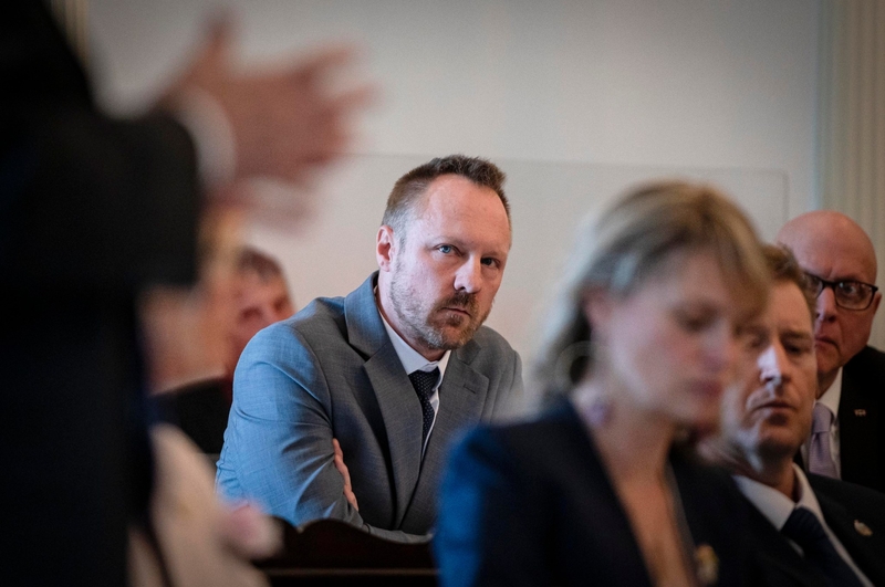 A man in a suit listens intently with arms crossed in a formal setting, surrounded by other seated individuals.