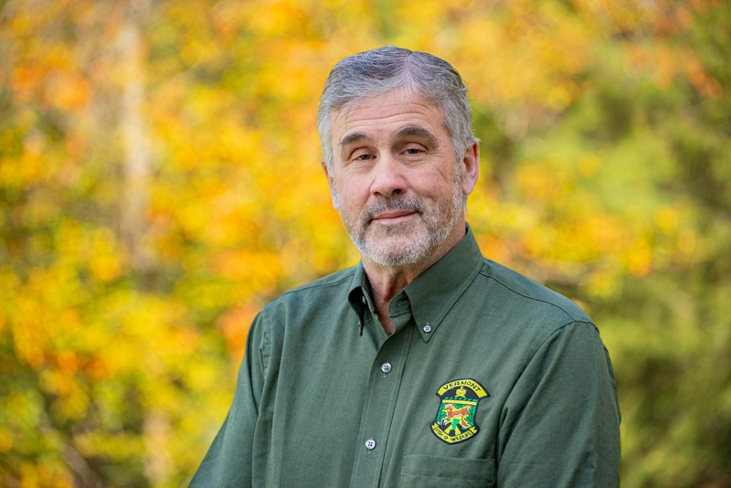 An older man with gray hair and a beard, wearing a green shirt with a badge, smiling gently against a blurred autumnal background.