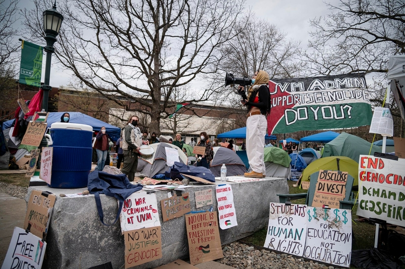 A person with a megaphone stands on concrete blocks at a busy outdoor protest with various signs and banners about human rights and divestment.
