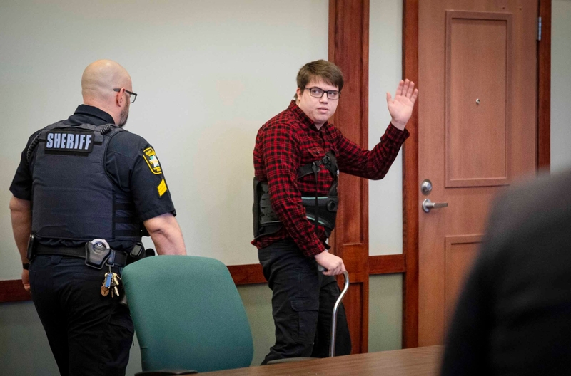 A person in a plaid shirt raising their hand in a courtroom as a sheriff's deputy looks on.