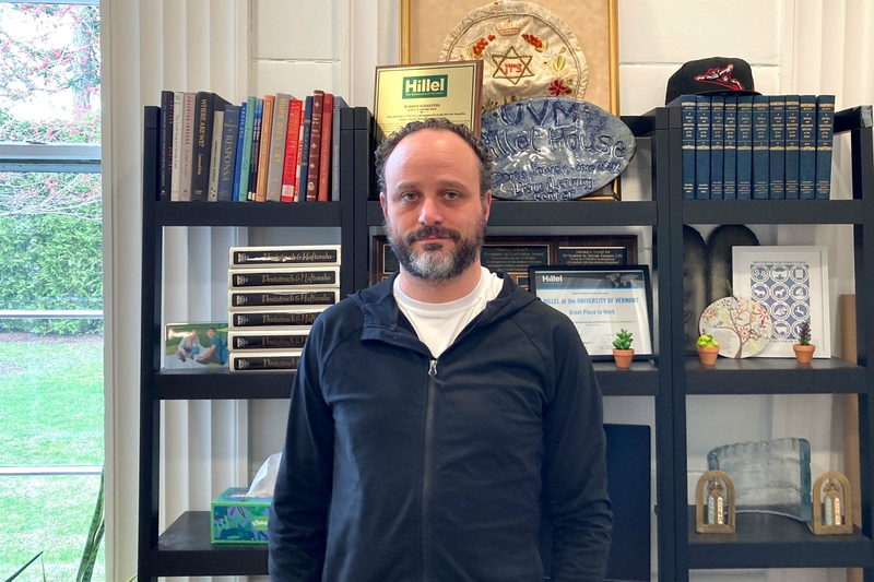 A man in a white shirt and black jacket stands in front of a bookshelf.
