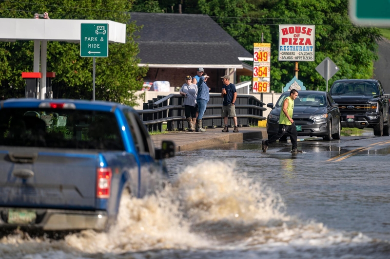 A flooded street with vehicles navigating the water. A person in a yellow jacket walks through the flood. A group of people stands on a nearby bridge. A "House of Pizza Restaurant" sign is visible.