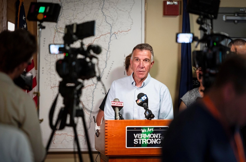 A man stands at a podium with microphones labeled WCAX, WPTZ, WE ARE VERMONT STRONG, addressing a group of people with cameras, against a backdrop of a map and an American flag.