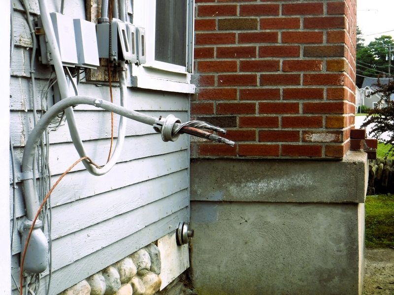 A damaged electrical cable sticks out from the side of a house, near a brick wall, with exposed wires.