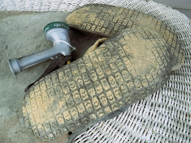 A worn pair of gardening knee pads with a textured surface lies next to a garden hose spray nozzle on a wicker surface.