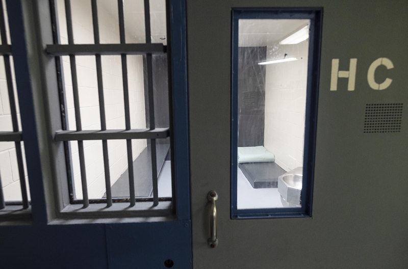 View of a jail cell with white walls, a grey door with a window labeled "HC," metal bars, a toilet, and a bed with a thin mattress.