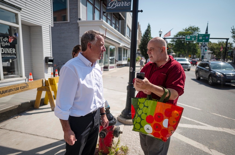 Two men are conversing on a street corner; one is holding a colorful shopping bag and a smartphone. A barricade, storefronts, and cars are visible in the background under a clear sky.
