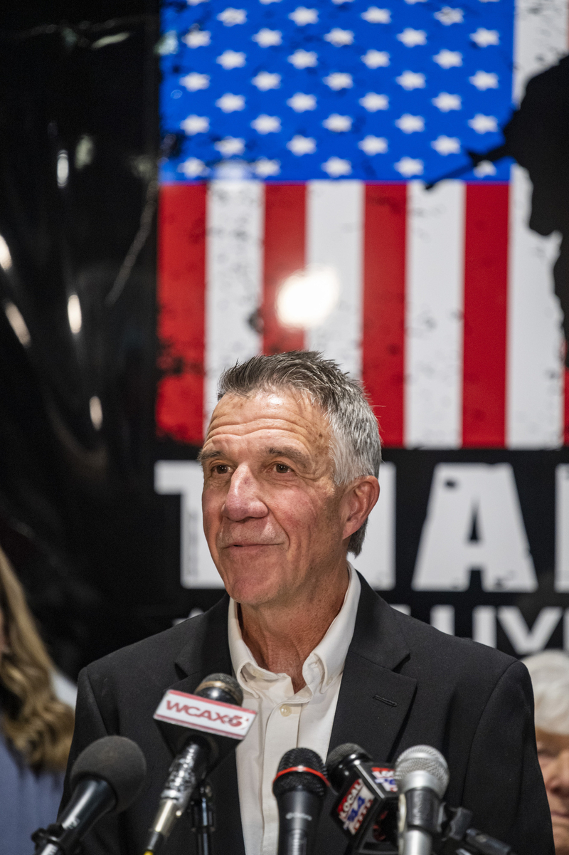 A man in a suit speaks at a podium with multiple microphones. An American flag is visible in the background.
