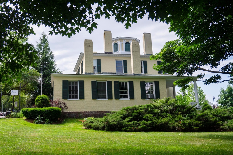 A large, two-story yellow house with green shutters, multiple chimneys, and a cupola, surrounded by green trees and shrubs on a well-kept lawn.