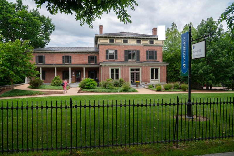A two-story brick building with shutters and a fenced lawn, featuring a sign labeled "Admissions" and "Taylor Hall." A person walks on a path in front.