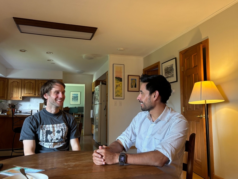 Two men sit across from each other at a wooden table in a well-lit kitchen, engaged in conversation. One person is wearing a t-shirt, the other a button-down shirt and smartwatch.