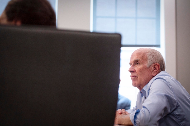 An older man in a blue shirt sits at a table, looking forward attentively during a meeting in a bright room.