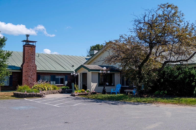 A single-story building with a green metal roof, brick chimney, and a small porch with blue chairs, next to a parking lot with a marked accessible parking space.