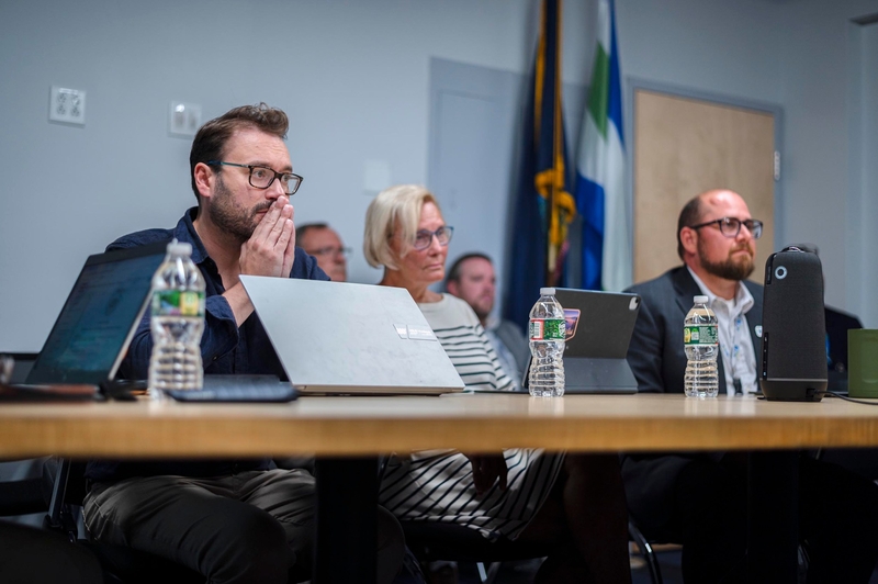 Several people sit at a conference table with laptops and water bottles, listening attentively during a meeting in an office setting.